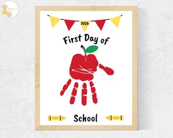 May include: A framed print featuring the text First Day of School with a red handprint shaped like an apple in the center. Above the text is a yellow and red bunting banner with the year 2026. Two yellow crayons flank the word School below.
