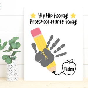 May include: A white printable sign with the text "Hip Hip Hooray! Preschool starts today!"  A handprint is shown holding a yellow pencil with a pink eraser. An apple with the name "Aiden" is drawn below the handprint.
