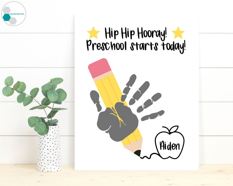 May include: A white printable sign with the text "Hip Hip Hooray! Preschool starts today!"  A handprint is shown holding a yellow pencil with a pink eraser. An apple with the name "Aiden" is drawn below the handprint.