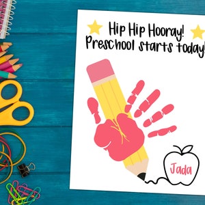 May include: A white card with the text "Hip Hip Hooray! Preschool starts today!" and a handprint in red paint holding a yellow pencil. The pencil is drawing an apple with the name "Jada" inside.