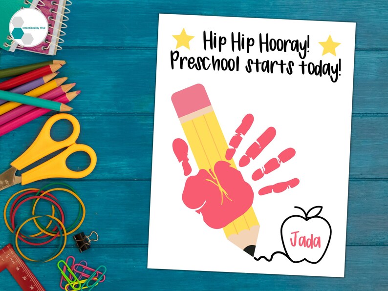May include: A white card with the text "Hip Hip Hooray! Preschool starts today!" and a handprint in red paint holding a yellow pencil. The pencil is drawing an apple with the name "Jada" inside.