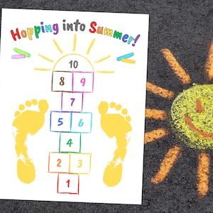 May include: A hopscotch game drawn on a white sheet of paper with yellow footprints and colorful squares numbered 1 to 10. The title "Hopping into Summer!" is written in rainbow colors. A yellow sun with a smiley face is drawn on a black asphalt surface.
