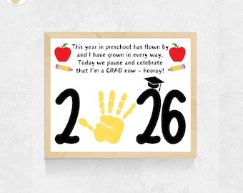 May include: A framed print with the text "This year in preschool has flown by...that I'm a GRAD now - hooray!" The print features the numbers "2" and "26", a yellow handprint, red apples, and pencils.