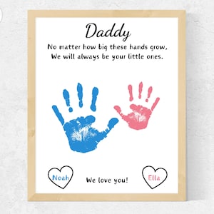 Könnte beinhalten: Ein gerahmter Druck mit einem Handabdruck-Design. Der Druck zeigt zwei Handabdrücke, einen blauen und einen rosa, mit dem Text "Daddy" oben. Der Text unter den Handabdrücken lautet "No matter how big these hands grow, We will always be your little ones." Der untere Teil des Drucks hat zwei Herzen mit den Namen "Noah" und "Ella" darin und den Text "We love you!" dazwischen.