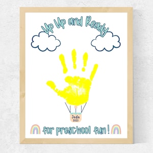 May include: A yellow handprint in the shape of a hot air balloon with the words "Up Up and Ready" above it. The balloon has a blue basket with the name "Jada" and the year "2023" written on it. Below the balloon are two rainbow arches and the words "for preschool fun!"