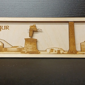 May include: A wooden plaque with a laser-cut design of a cityscape. The plaque is light brown and has the text "Ayan Baqur" at the top.