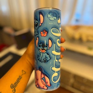 Puede incluir: Un vaso de acero inoxidable azul y rosa con un dise&ntilde;o de personaje de dibujos animados. El vaso presenta un personaje azul con orejas grandes y un personaje rosa con una nariz larga. El vaso est&aacute; decorado con flores y la palabra "Ohana".