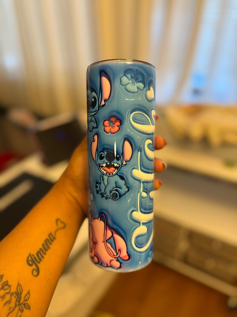 Puede incluir: Un vaso de acero inoxidable azul y rosa con un dise&ntilde;o de personaje de dibujos animados. El vaso presenta un personaje azul con orejas grandes y un personaje rosa con una nariz larga. El vaso est&aacute; decorado con flores y la palabra "Ohana".