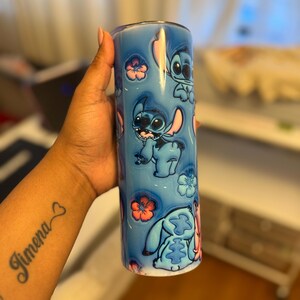 Puede incluir: Un vaso de acero inoxidable azul con tapa blanca y un dise&ntilde;o de Stitch de Lilo & Stitch. El dise&ntilde;o presenta a Stitch en varias poses con flores rosas.