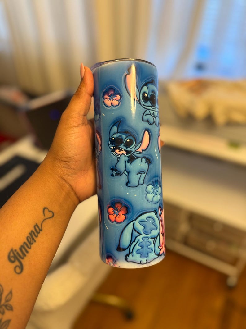 Puede incluir: Un vaso de acero inoxidable azul con tapa blanca y un dise&ntilde;o de Stitch de Lilo & Stitch. El dise&ntilde;o presenta a Stitch en varias poses con flores rosas.