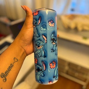 Puede incluir: Un vaso de acero inoxidable azul y blanco con un patr&oacute;n repetido de personajes de Stitch de la pel&iacute;cula de Disney Lilo & Stitch. El vaso tiene una tapa transparente y est&aacute; decorado con flores rosas.