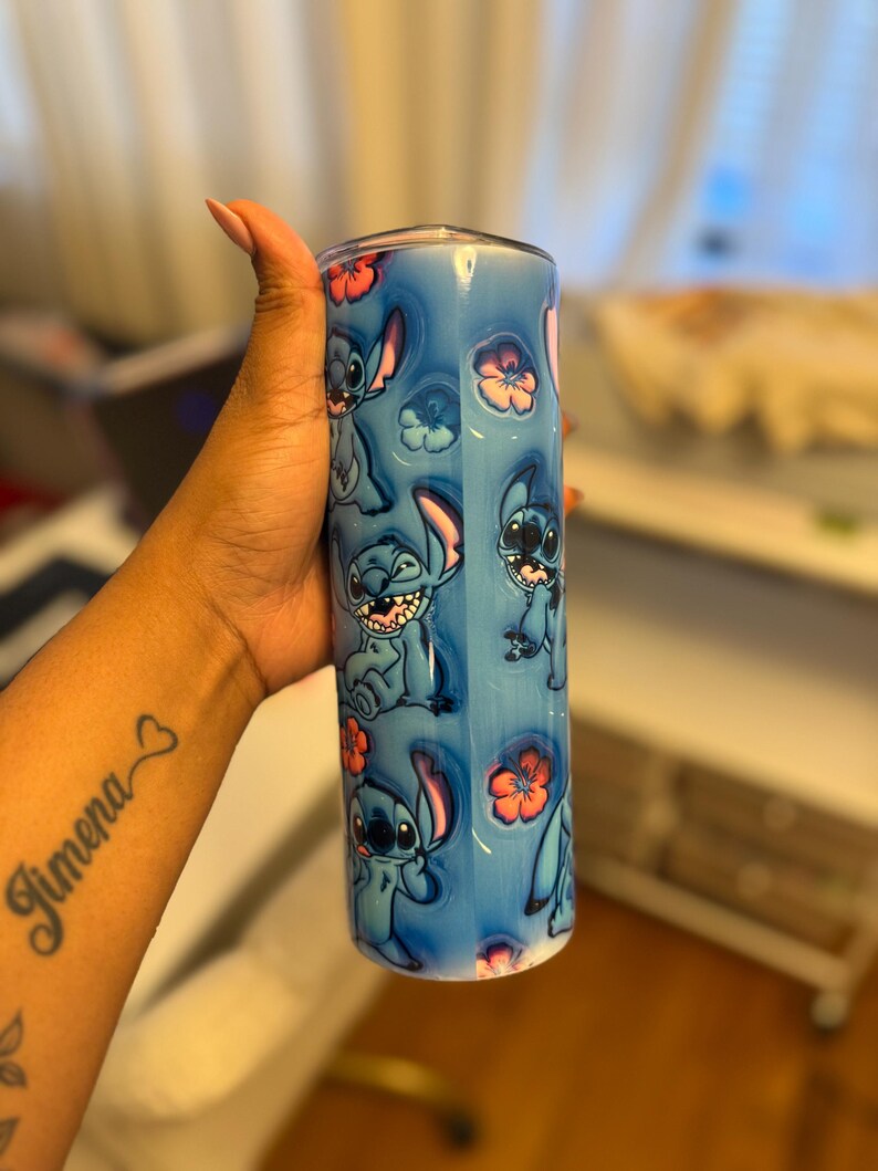 Puede incluir: Un vaso de acero inoxidable azul y blanco con un patr&oacute;n repetido de personajes de Stitch de la pel&iacute;cula de Disney Lilo & Stitch. El vaso tiene una tapa transparente y est&aacute; decorado con flores rosas.