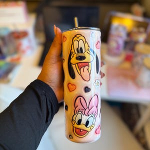 May include: A pink and white stainless steel tumbler with a straw. The tumbler features a cartoon image of Pluto and Daisy Duck with hearts and other designs.