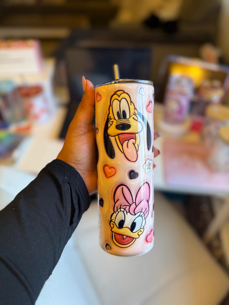 May include: A pink and white stainless steel tumbler with a straw. The tumbler features a cartoon image of Pluto and Daisy Duck with hearts and other designs.