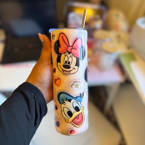 May include: A white stainless steel tumbler with a pink and blue cartoon design featuring Mickey Mouse and Donald Duck. The tumbler has a straw and is decorated with hearts and flowers.