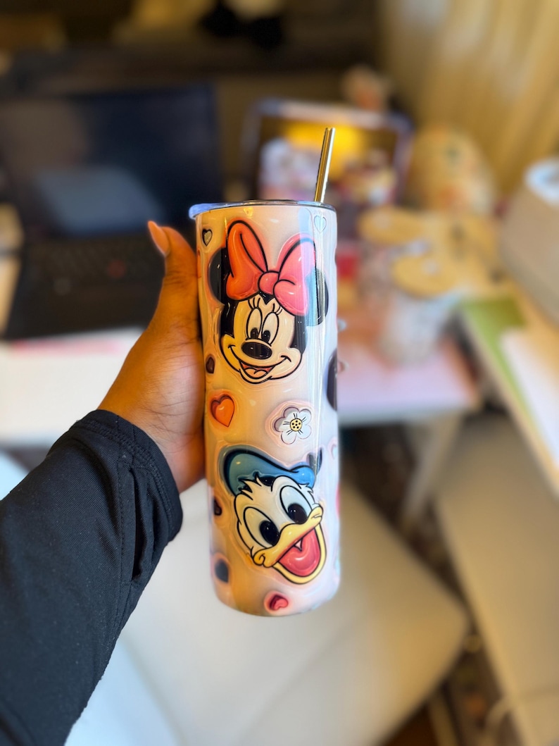 May include: A white stainless steel tumbler with a pink and blue cartoon design featuring Mickey Mouse and Donald Duck. The tumbler has a straw and is decorated with hearts and flowers.