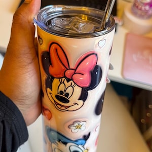 May include: A pink and white stainless steel tumbler with a straw. The tumbler features a design of Minnie Mouse and Donald Duck with hearts and flowers.