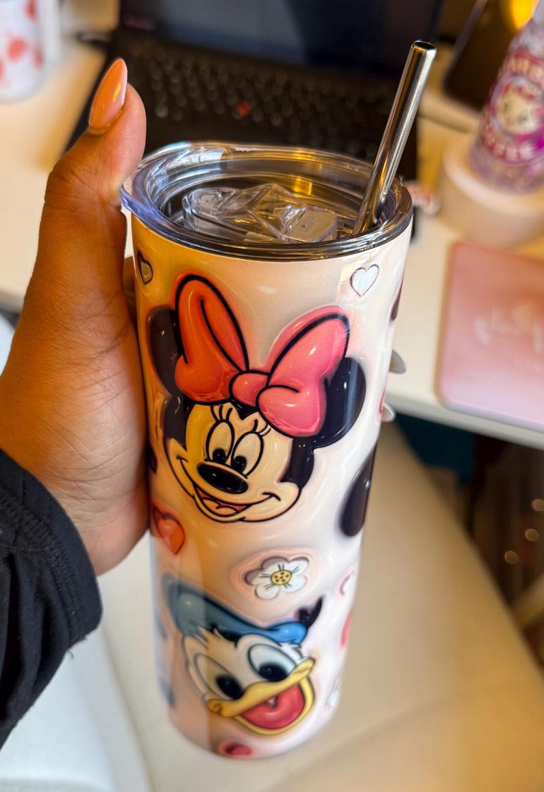 May include: A pink and white stainless steel tumbler with a straw. The tumbler features a design of Minnie Mouse and Donald Duck with hearts and flowers.
