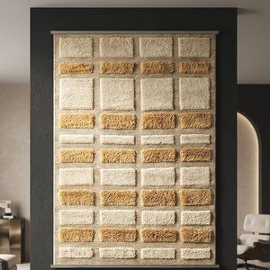 Könnte beinhalten: Ein rechteckiger Wandbehang mit einem strukturierten, ziegelartigen Design. Das Muster wechselt zwischen beigen und goldbraunen rechteckigen Blöcken und erzeugt einen dreidimensionalen Effekt. Das Stück ist auf einem neutralen Hintergrund montiert.