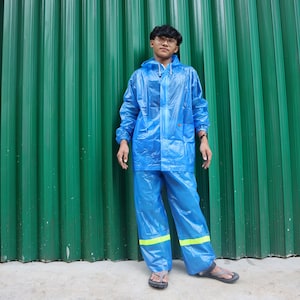May include: A person wearing a blue raincoat with a yellow stripe on the legs. The raincoat is made of a shiny, waterproof material. The person is standing in front of a green corrugated metal wall.