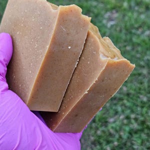 May include: Two rectangular bars of solid, light brown soap with a textured, speckled surface. The soap bars have a matte finish and appear firm, with visible small particles embedded throughout the light tan, earthy-toned material.