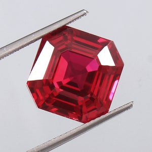 May include: A faceted, octagonal-cut ruby gemstone held by tweezers. The gem is a deep, saturated red colour with sharp, defined facets that reflect light. The gemstone is a brilliant, eye-catching piece.