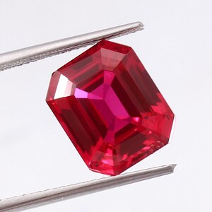 May include: A faceted, rectangular-cut ruby gemstone held by silver tweezers. The ruby is a vibrant red color with a high degree of clarity and brilliance. The gemstone's facets reflect light, creating a sparkling effect. The tweezers provide a contrasting metallic element.
