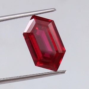 Op de afbeelding: Een gefacetteerde, rechthoekige robijn edelsteen, vastgehouden door een pincet. De edelsteen heeft een dieprode kleur en een geometrische slijpvorm met meerdere facetten. De pincet is zilverkleurig en de achtergrond is lichtgrijs.