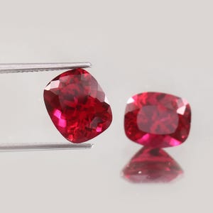 Puede incluir: Dos piedras preciosas de color rojo intenso con corte cojín y superficies facetadas que reflejan la luz. Una piedra es sostenida por unas pinzas de metal plateado, mientras que la otra descansa cerca, mostrando un reflejo claro en la superficie.