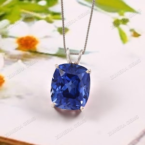 Op de afbeelding: Een zilveren hangerketting met een grote, kussenvormige, diepblauwe edelsteen. De edelsteen is gezet in een zilveren zetting en hangt aan een fijne zilveren ketting. De achtergrond is een zacht, wazig bloemenpatroon.