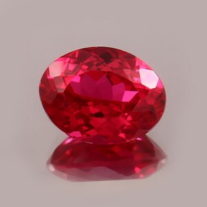May include: A faceted, oval-cut ruby gemstone. The gem is a vibrant red colour, with multiple facets reflecting light. The ruby is set against a light background, with its reflection visible below.
