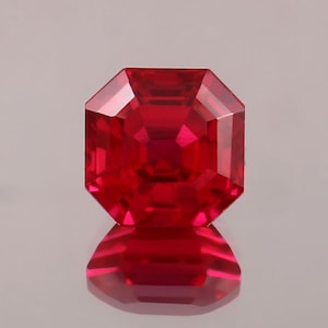 May include: A faceted, square-cut ruby gemstone. The gem is a deep, vibrant red color with a reflective surface. The gemstone is displayed on a reflective surface, creating a mirror image of the stone.