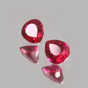 May include: Two vibrant, deep red, pear-shaped faceted gemstones. Each stone features a pointed end and a rounded top, with precise geometric cuts that create internal light reflections and a brilliant, polished surface appearance.
