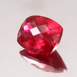 May include: A faceted, cushion-cut gemstone in a vibrant shade of red. The gem's surface reflects light, creating a sparkling effect. The stone is displayed on a reflective surface, enhancing its brilliance. This gemstone is likely for jewelry.