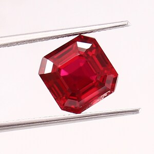 Puede incluir: Un rubí tallado en forma cuadrada, sostenido por unas pinzas. El rubí es de un color rojo intenso y saturado, con un brillo brillante. Las facetas de la gema reflejan la luz, creando un efecto brillante. Las pinzas son de color plateado.