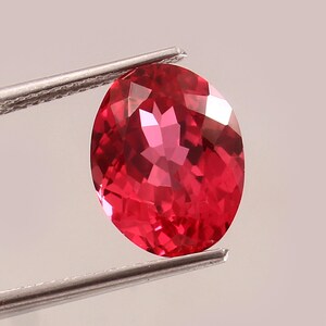 Peut inclure: Une pierre précieuse rouge, de forme ovale et facettée, est maintenue par une pince argentée. La surface polie de la gemme reflète la lumière, révélant sa structure interne. La couleur est un rouge vif et saturé.