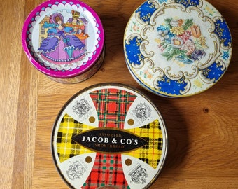 May include: Three round tin boxes with different designs. The first box has a pink background with a colorful illustration of a woman in a blue dress and a man in a red suit. The second box has a blue background with a white floral design. The third box has a red and black plaid design with the text "Assorted Jacob & Co's Shortbread".
