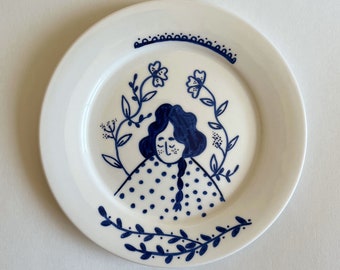 以下が含まれることがあります： 青い手描きのデザインが施された白いセラミックプレート。デザインは、青い髪と青い水玉模様のドレスを着た人物が、青い花と葉に囲まれている様子を表しています。