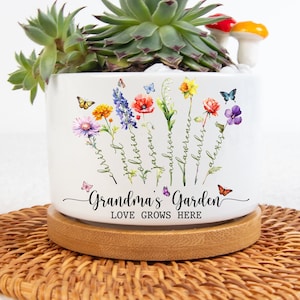 May include: White ceramic planter with a floral design and the text "Grandma's Garden Love Grows Here". The planter has a wooden base and is filled with succulents and a small mushroom figurine.
