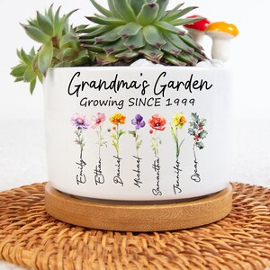 May include: White ceramic planter with the text "Grandma's Garden Growing SINCE 1999" and a floral design with names written below each flower. The planter is sitting on a woven brown coaster.