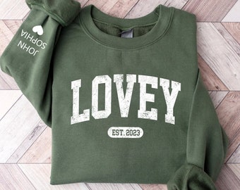 May include: Green sweatshirt with the word "LOVEY" in white distressed lettering and the text "EST. 2023" in a white oval below. The sweatshirt has a white heart and the names "JOHN" and "SOPHIA" on the left sleeve.