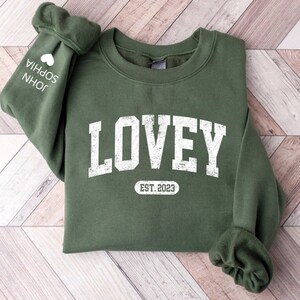 May include: Green sweatshirt with the word "LOVEY" in white distressed lettering and the text "EST. 2023" in a white oval below. The sweatshirt has a white heart and the names "JOHN" and "SOPHIA" on the left sleeve.