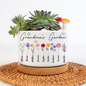 May include: White ceramic planter with the text "Grandma's Garden" and a row of flowers with names written below each flower. The names are Ethan, Bean, Miller, Jimmie, Sophia, Joshua, Graham, Emma, and Amelia. The planter is sitting on a wooden coaster.
