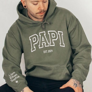 May include: A green hooded sweatshirt with the word "PAPI" in white outlined letters and the text "EST. 2024" below. The sweatshirt has a small heart and the names "Liam", "Ethan", and "Sophia" on the left sleeve.