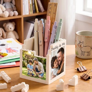 May include: A white square desk organizer constructed from interlocking building blocks, featuring two photo panels showing a father and child. The organizer holds several pens and a wooden ruler marked up to 30 centimeters in length.