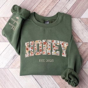 May include: A green sweatshirt with the word "HONEY" in floral print letters. The text "EST. 2025" is printed below the word "HONEY".