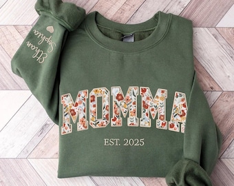 Op de afbeelding: Een groen sweatshirt met het woord "MOMMA" in bloemenprint en de tekst "EST. 2025" eronder. Het sweatshirt heeft een gouden hart met de tekst "Ellie" en "Sophie" op de linkermouw.