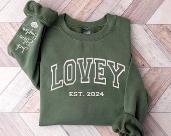 May include: A green sweatshirt with the word "LOVEY" in white with a white outline, and the text "EST. 2024" below it. The sweatshirt has a white heart and the text "Jack, Ethan, Hudson" on the left sleeve.