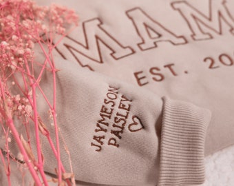 Puede incluir: Una sudadera de color marrón claro con la palabra "MAM" en letras grandes de color marrón y el texto "EST. 20" debajo. La manga tiene el texto "JAYMESON PAISLEY" y un corazón.
