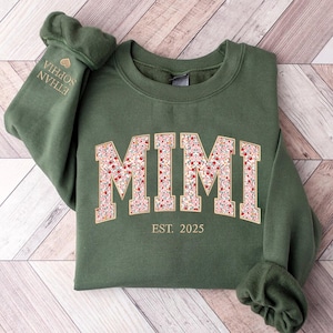 May include: Green sweatshirt with the word "MIMI" in a floral pattern across the chest. The text "EST. 2025" is below the name. The left sleeve has the names "ETHAN SOPHIA" embroidered in gold.